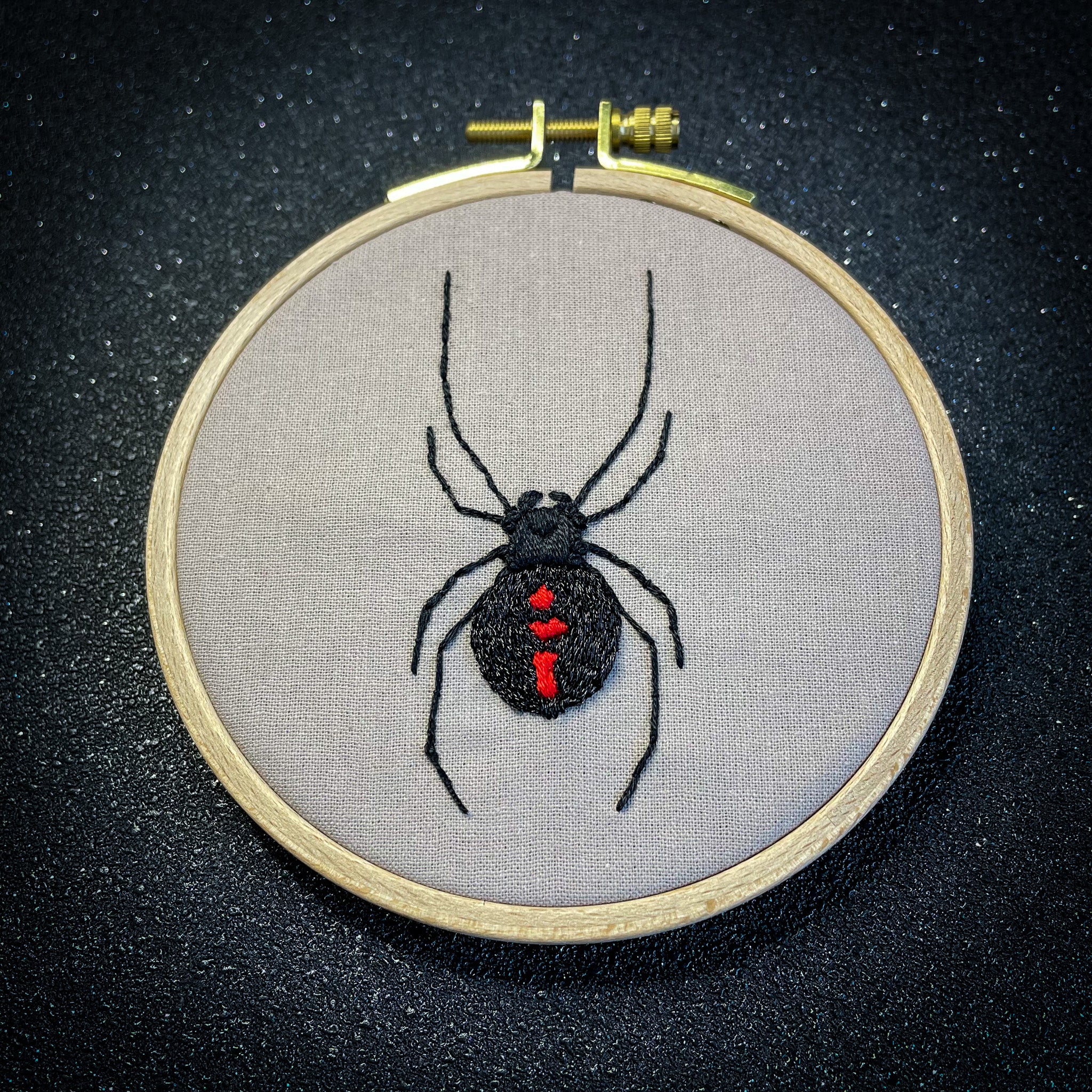 Image shows a sparkly black background. In the foreground is an embroidery hoop containing a stitched design of a black widow spider. This is an image of a finished embroidery kit design. The design is stitched on sand coloured fabric. The spider is stitched in black metallic thread with red markings on the spider's body.