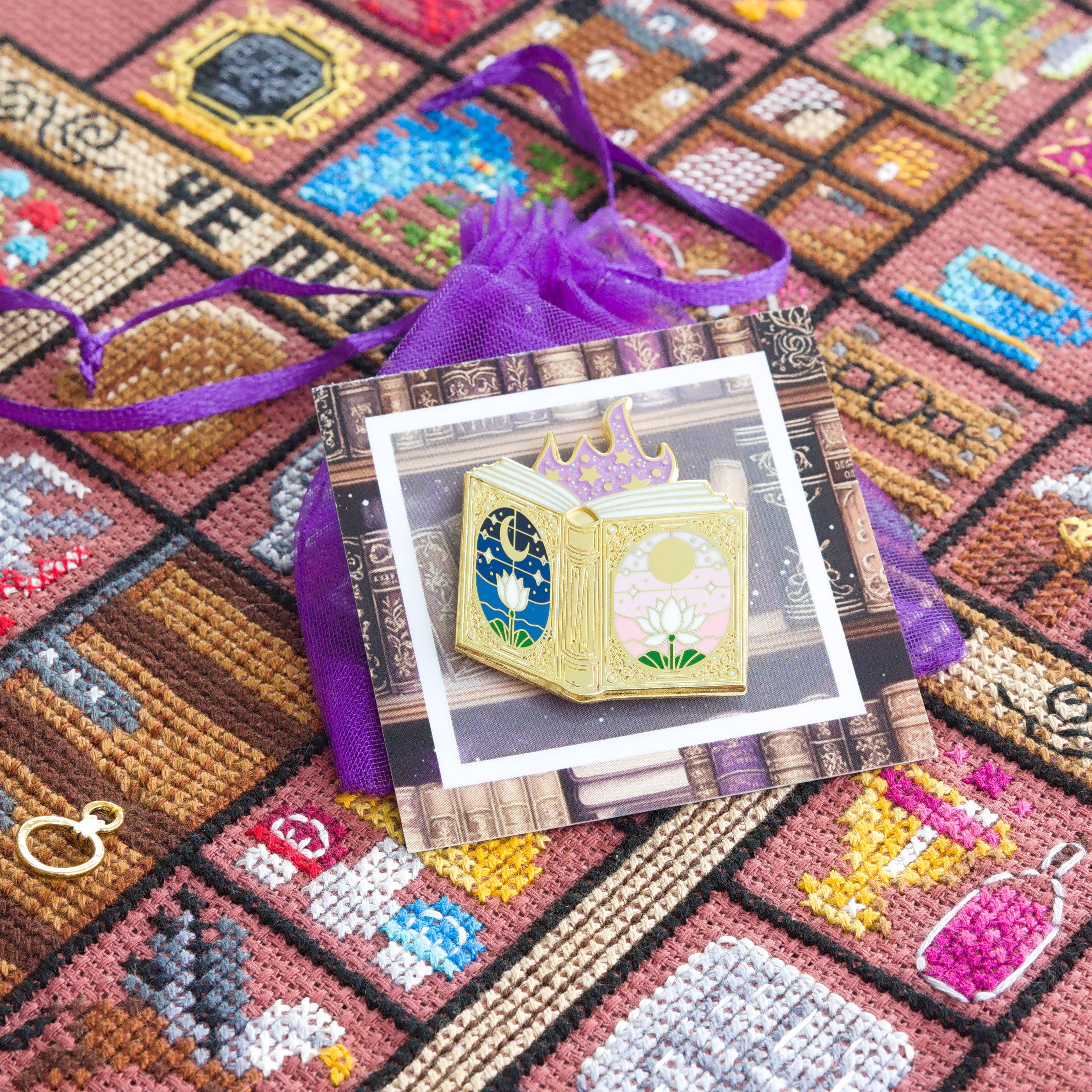Needle minder shaped like a fairytale book, attached to a backing card with a design showing old books, resting on a purple organza bag.