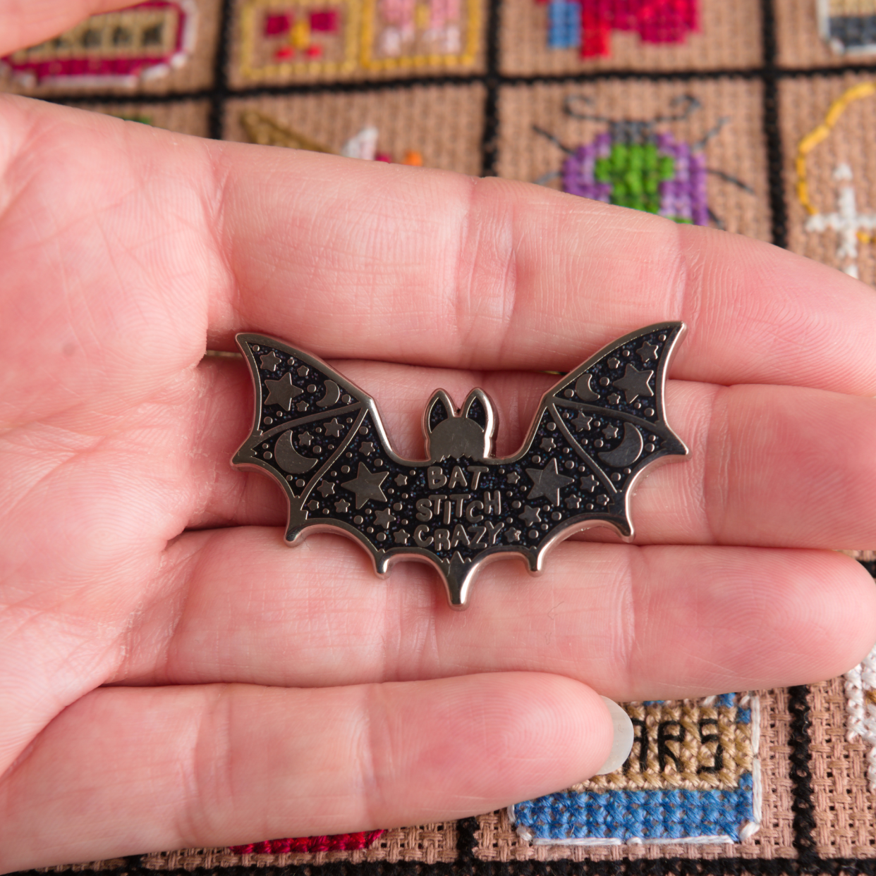 Hand holding a bat-shaped needle minder with a cross-stitch background