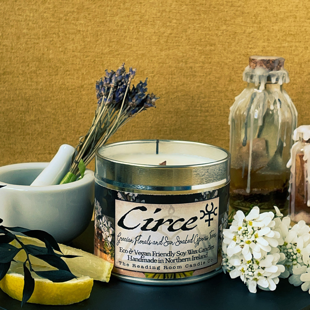 Handmade soy candle in a tin. The scent is lavender, cyclamen and cypress trees. The scent is refreshing but calming. The image shows the candle called 'Circe' surrounded by items associated with witches. Potion bottles, herbs, lavender, lemons and summer flowers are around the candle.