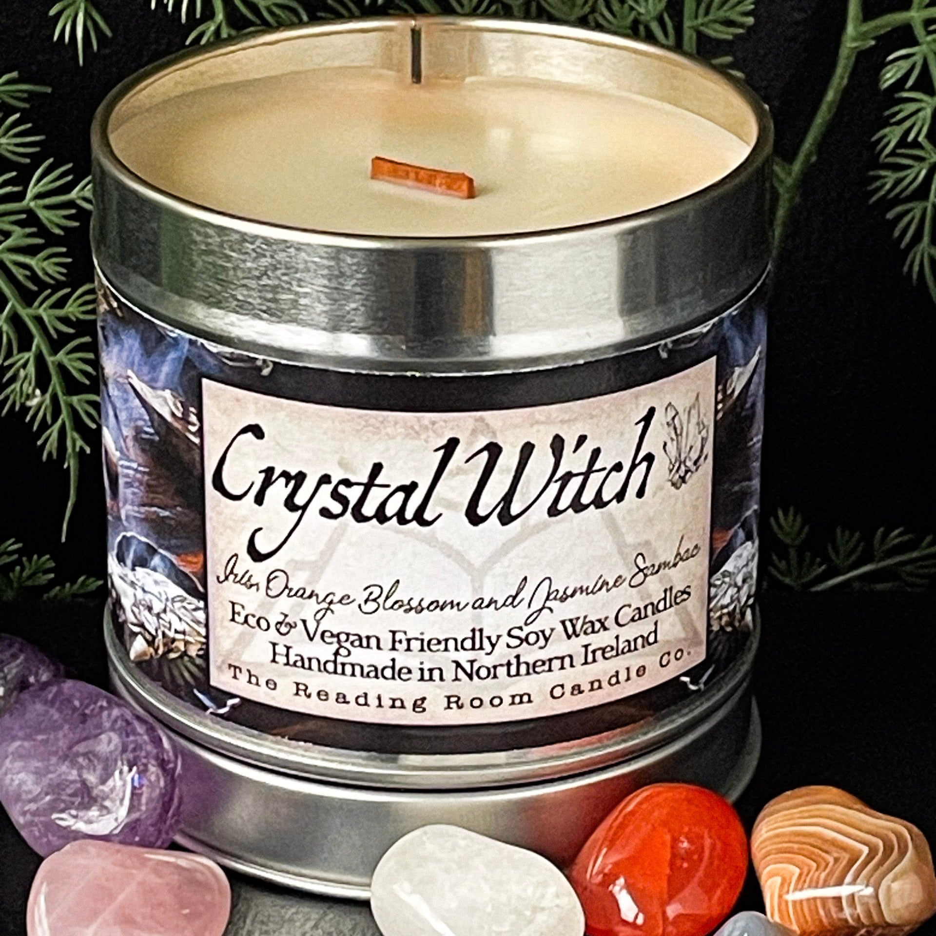 Candle in a metal container labeled 'Crystal Witch' surrounded by crystals and leaves.