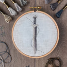 Image shows a light wood table top on which is a finished embroidery design of a fantasy style sword. Stitched in metallic silvers and greys on an ice blue fabric. On the table top around the embroidery in a hoop, are thread skeins used in the design, a needle, scissors and decorative shiny snowflakes.