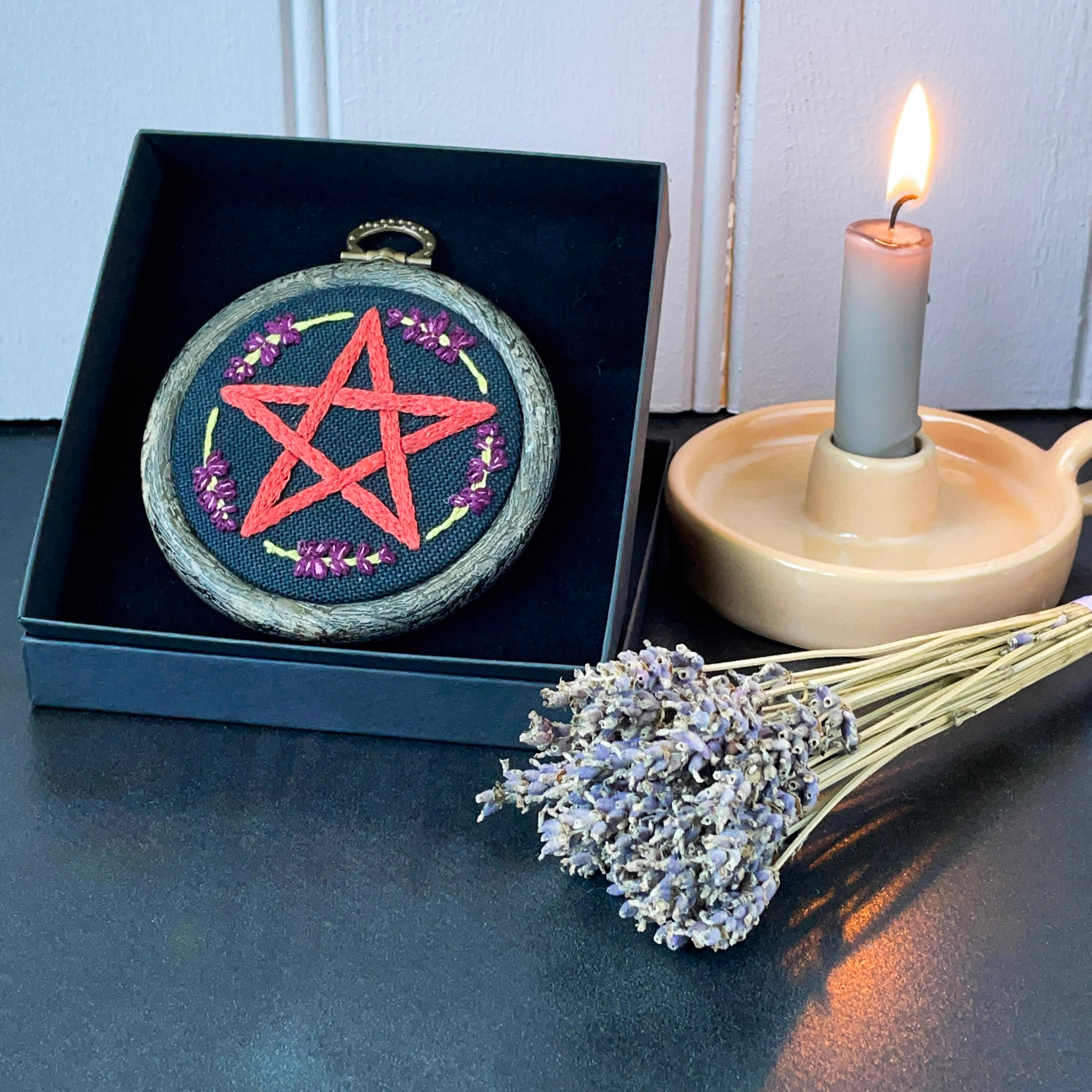 Embroidery hoop with a pentagram design on a dark surface next to a candle and lavender flowers.