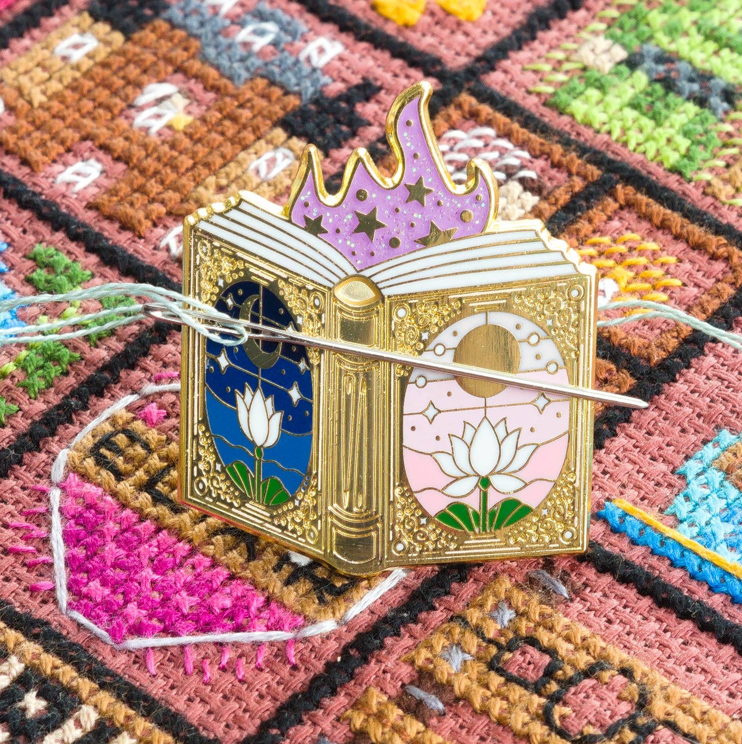 Needle minder shaped like a fairytale book, with gold and lotus flowers. Image shows the needle minder with a needle, against a cross stitch background.