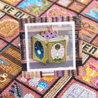 Close-up of a fairy tale book needle minder on a backing card with a book design. The card and needle minder are displayed against cross-stitched pattern background.
