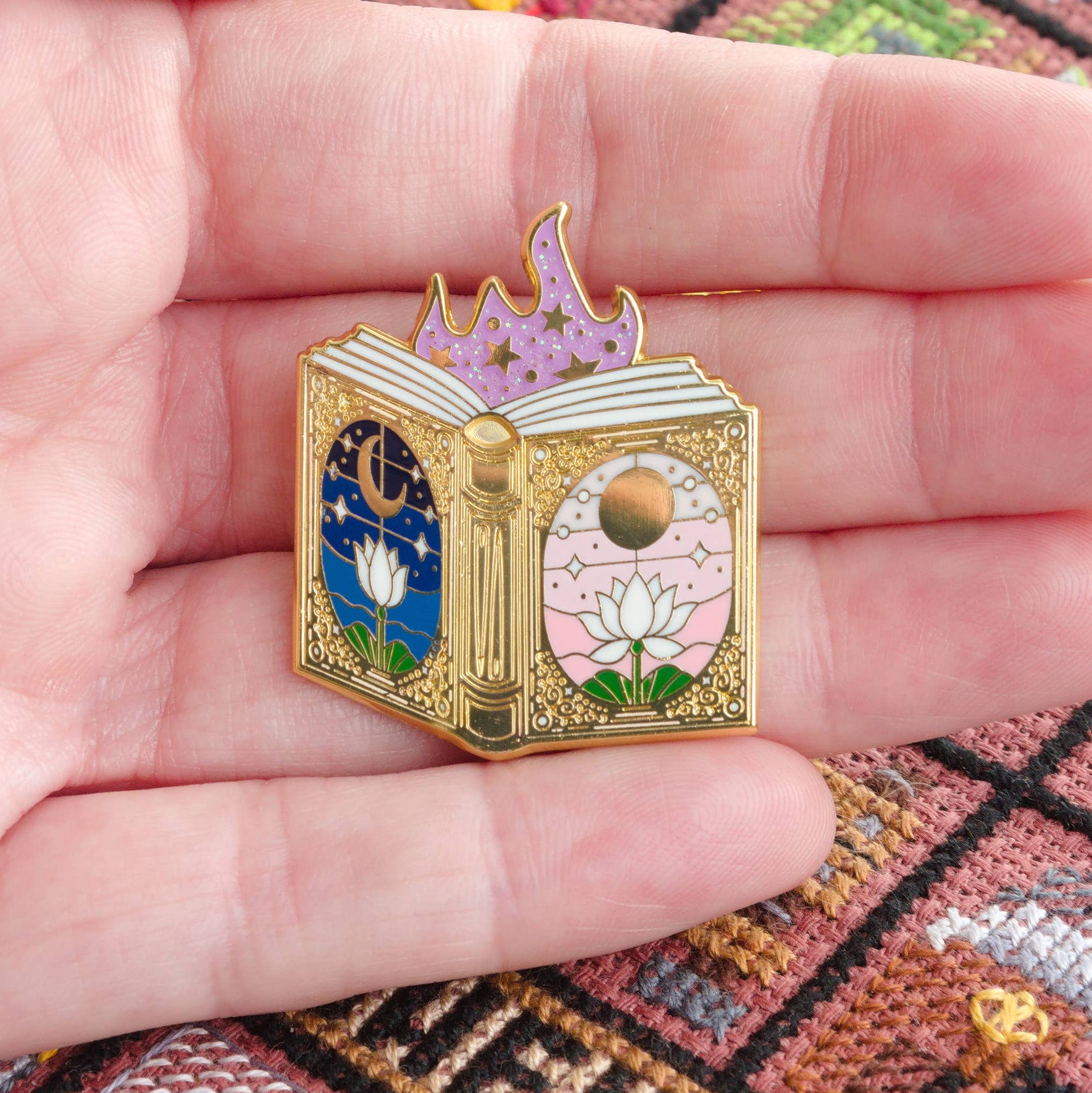 Hand holding a small, intricately designed book-shaped needle minder against a cross-stitched background.