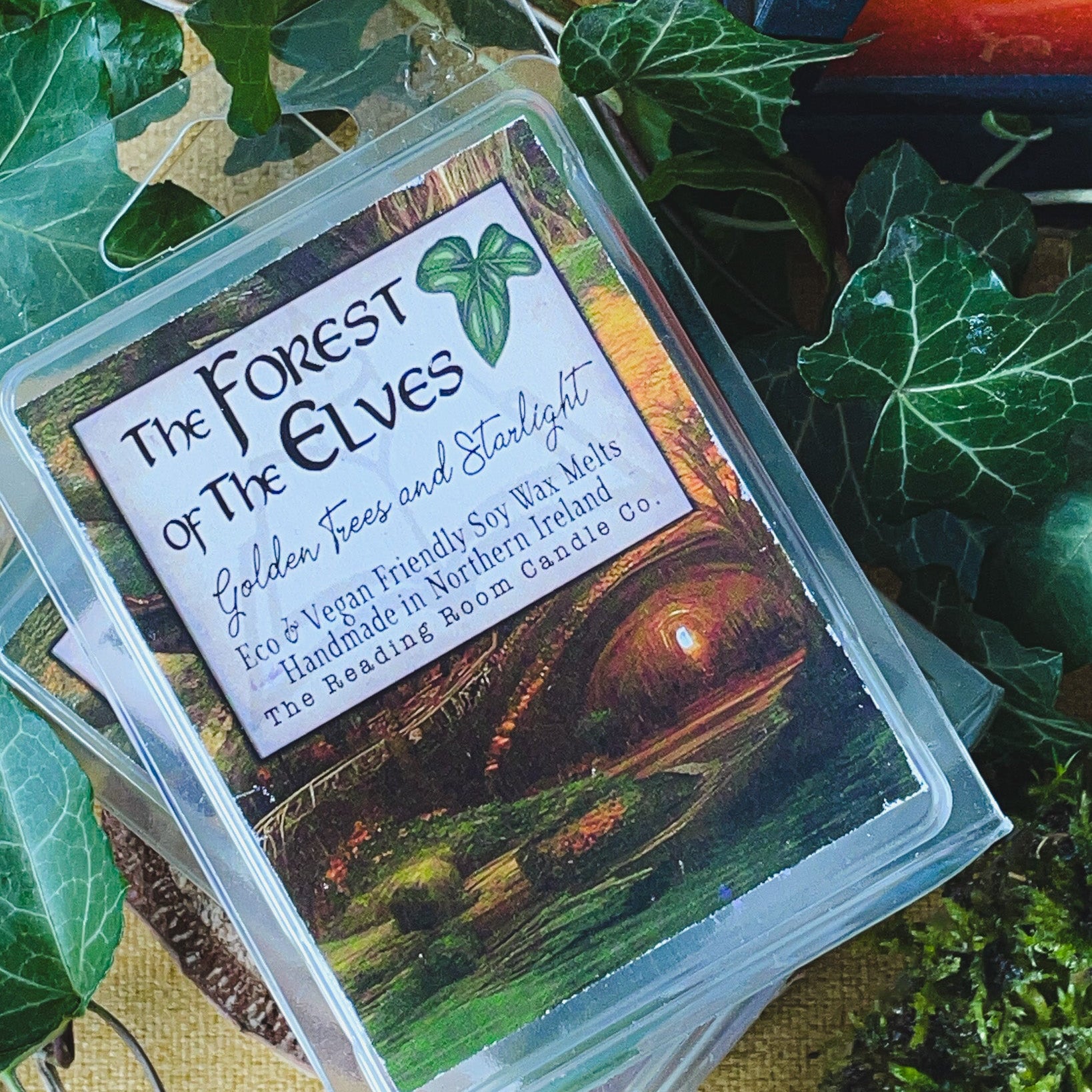 A soy wax melt pack called 'The Elves of the Forest' with an illustration of a sunlit forest scene, placed on a table with green leaves and moss.