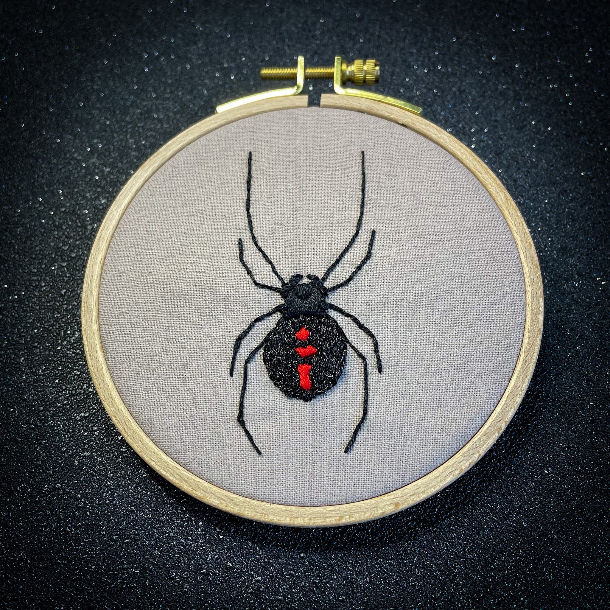 Image shows a sparkly black background. In the foreground is an embroidery hoop containing a stitched design of a black widow spider. This is an image of a finished embroidery kit design. The design is stitched on sand coloured fabric. The spider is stitched in black metallic thread with red markings on the spider's body. 