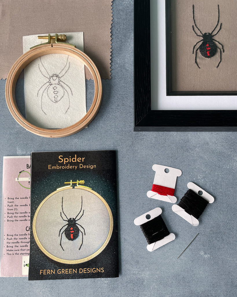 A flat lay image of embroidery kit contents for a spider design. The image shows the completed spider design in one corner and the kit contents spaced neatly to the left and bottom of the finished design. The contents shown are instruction sheet, stitch guide, needle, thread, a hoop, fabric and a pre-drawn spider design.