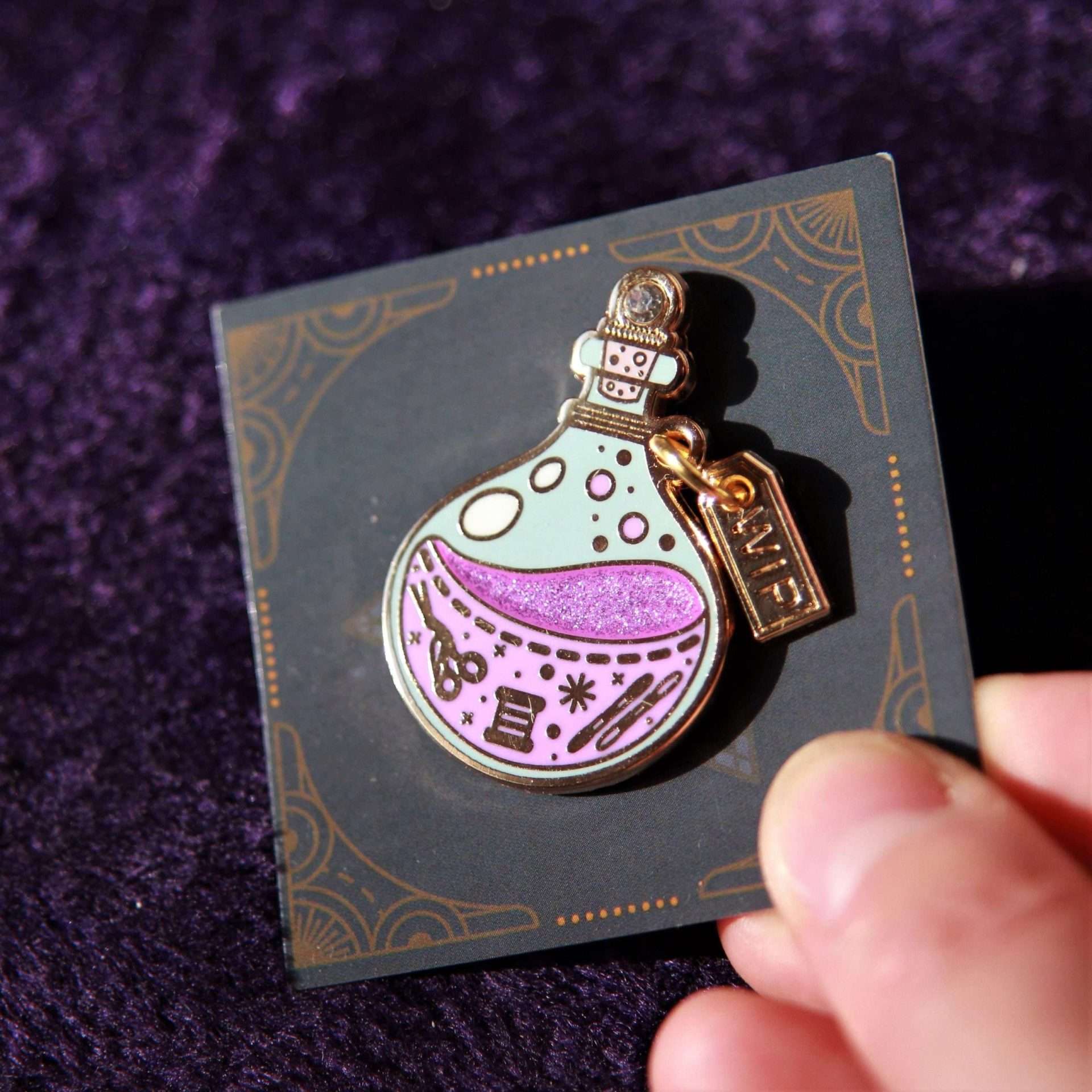 Enamel needle minder of a potion bottle with a 'WIP' tag on a black card, held against a purple fabric background.