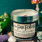 A tin candle called 'The Fair Folk', with the scent of citrus cassis, jasmine and violet. Around the base of the tin are crystals and rocks, one is a pale purple. In the background is a red and white spotted fly agaric mushroom and some green leaves.