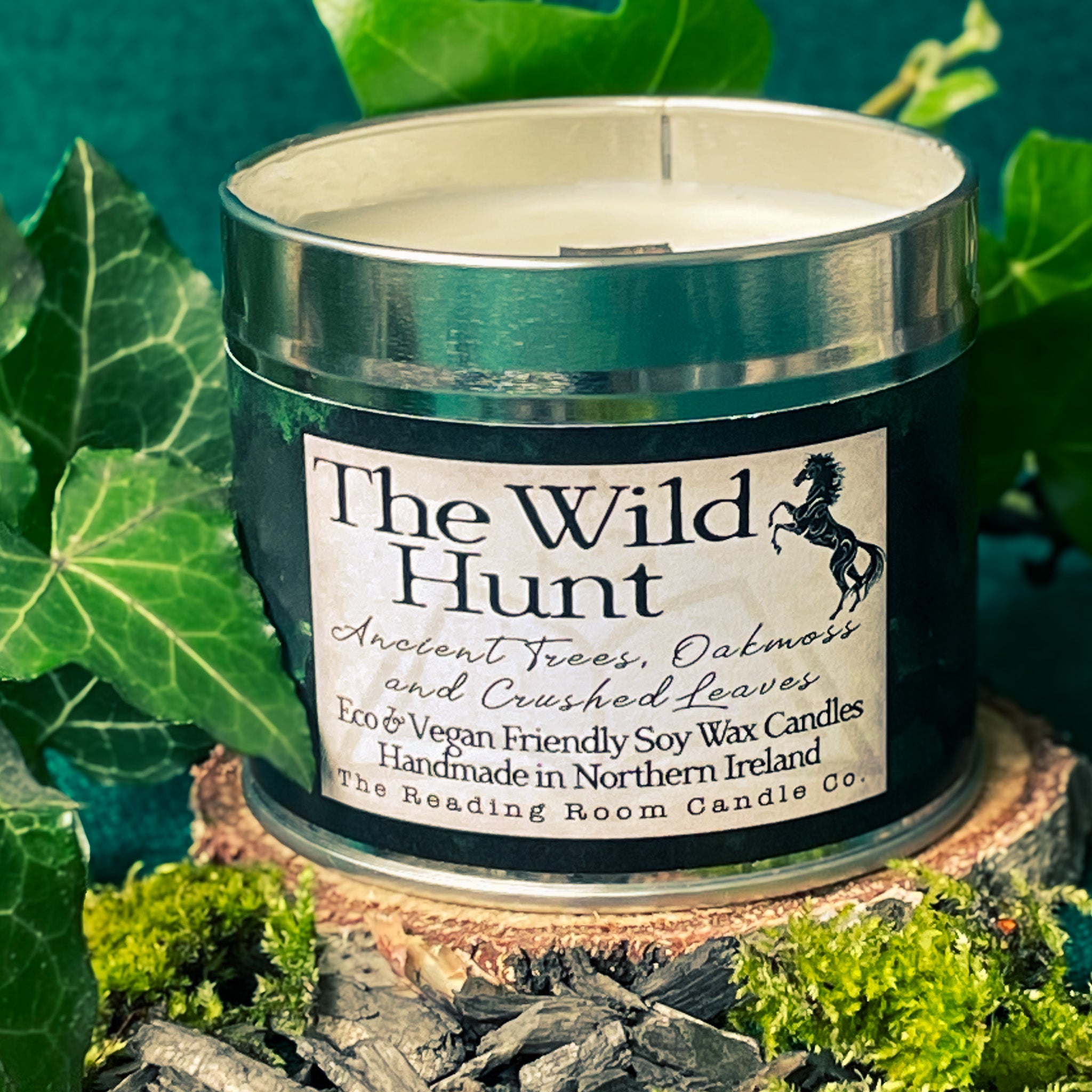 Tin candle called 'The Wild Hunt' is in the foreground, surrounded by ivy leaves, moss and sitting atop a small slice of tree trunk.