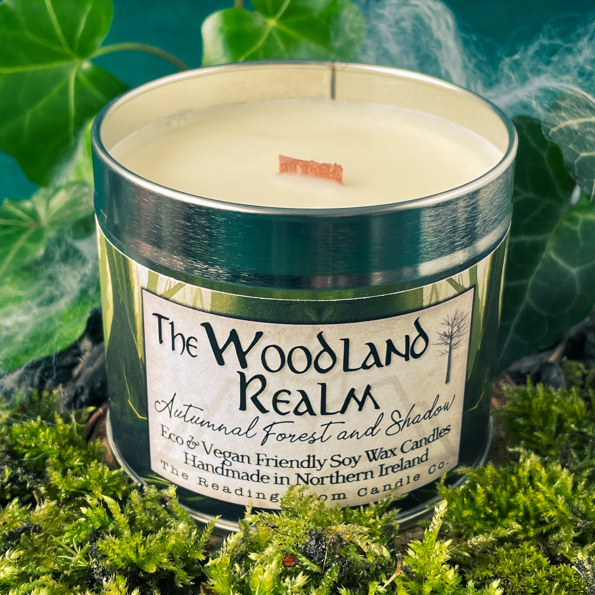 Tin candle called 'The Woodland Realm' sitting on top of a mound of moss. Ivy leaves around the back of the tin and a dark blue background. Mist swirls around the leaves to the right of the picture.