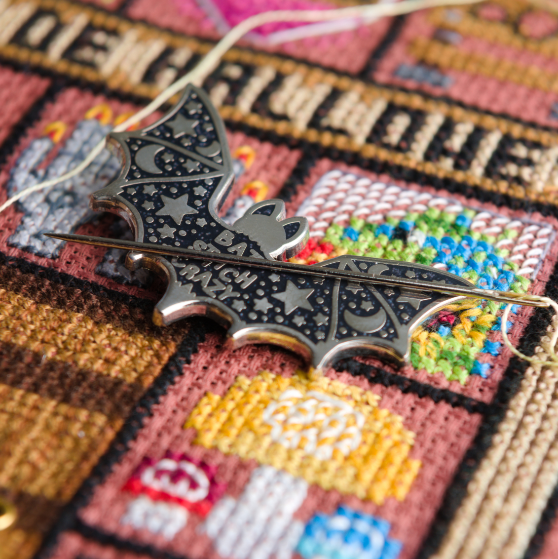 Silver and black bat shaped needle minder with needle on a cross-stitched background.