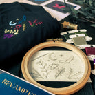 Image taken from above angle shows contents of embroidery kit designed for revamping clothes. T-shirt with completed design, embroidery hoop, thread and instruction sheets can be seen. The embroidery design is a mystical theme with stars, moon, bats, pentagrams, flowers and leaves.